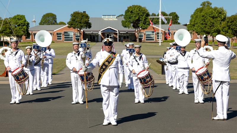 Navy band members in white uniform stand in a line formation while playing instruments.