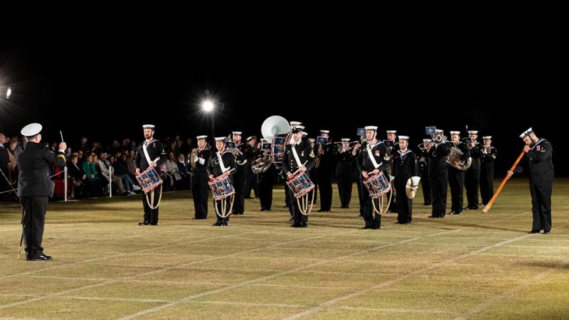 Navy Band musicians play in lined formation on a field at night in front of a seated audience.