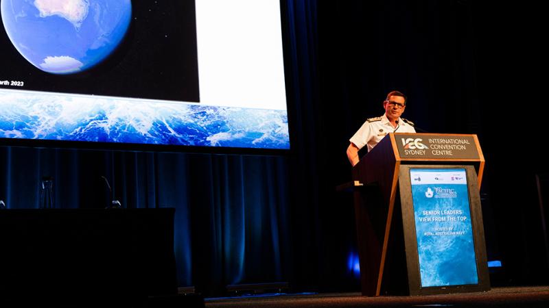 A speaker in white Navy uniform stands behind a podium in front of a large screen showing an image of earth from space.
