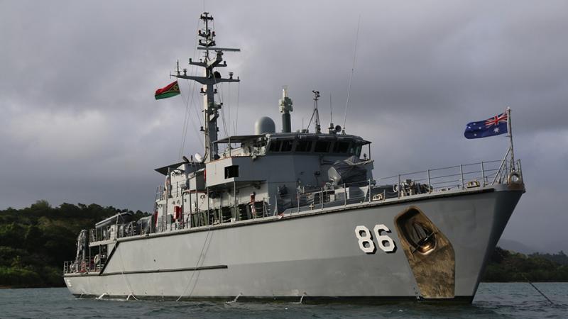 HMAS Diamantina anchored off a tree-lined coast on an overcast day.