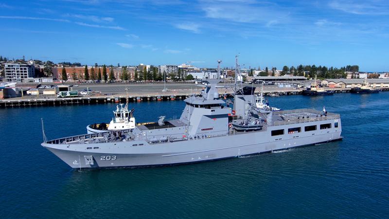 A large grey vessel sails in front of a shipping terminal on a clear day.