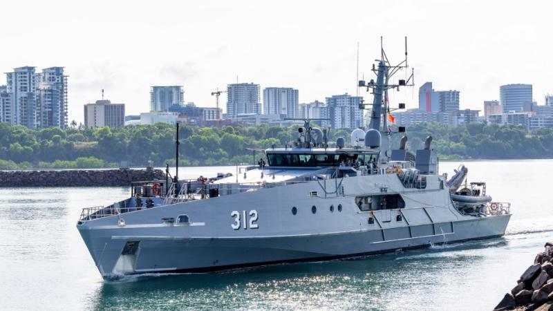 A small grey ship in calm river waters on an overcast day with low set city buildings in the background.