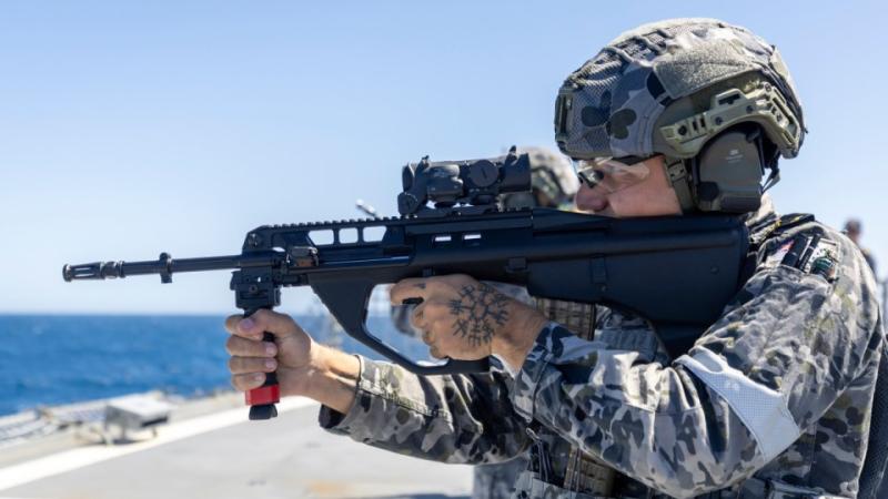 A person wearing a camouflage uniform, helmet, protective eyewear and headset holds a rifle while standing on a ship, with the ocean visible in the background.